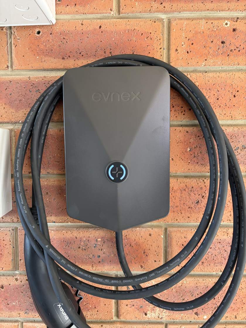 An Evnex EV charger installed by East Arc Group on the red brick wall of an Adelaide house, powered up with its indicator ring lit blue and the charging lead coiled neatly around the body of the unit