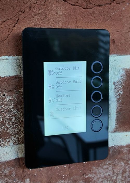 A KASTA e-ink scene controller mounted on a rustic red brick wall, its display listing four named scenes: Outdoor DLs off, Outdoor Wall off, Heaters, and Outdoor Chill, with a page indicator showing one of four pages