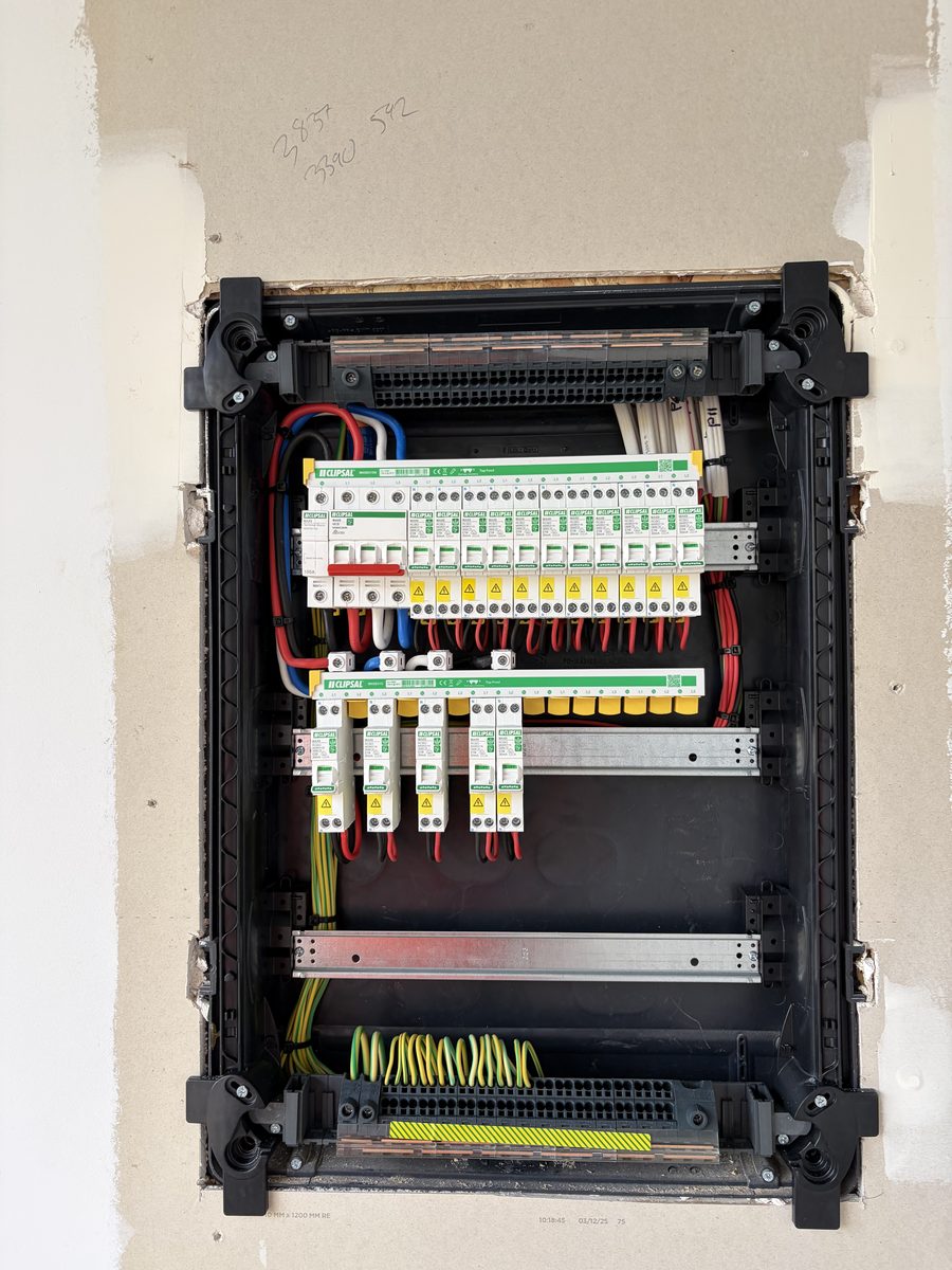 An East Arc Group switchboard enclosure recessed into an unfinished stud wall, the drywall cut edges still raw around it, with the main switch and a full run of Clipsal RCBOs already fitted and wired but no cover on and no labels yet