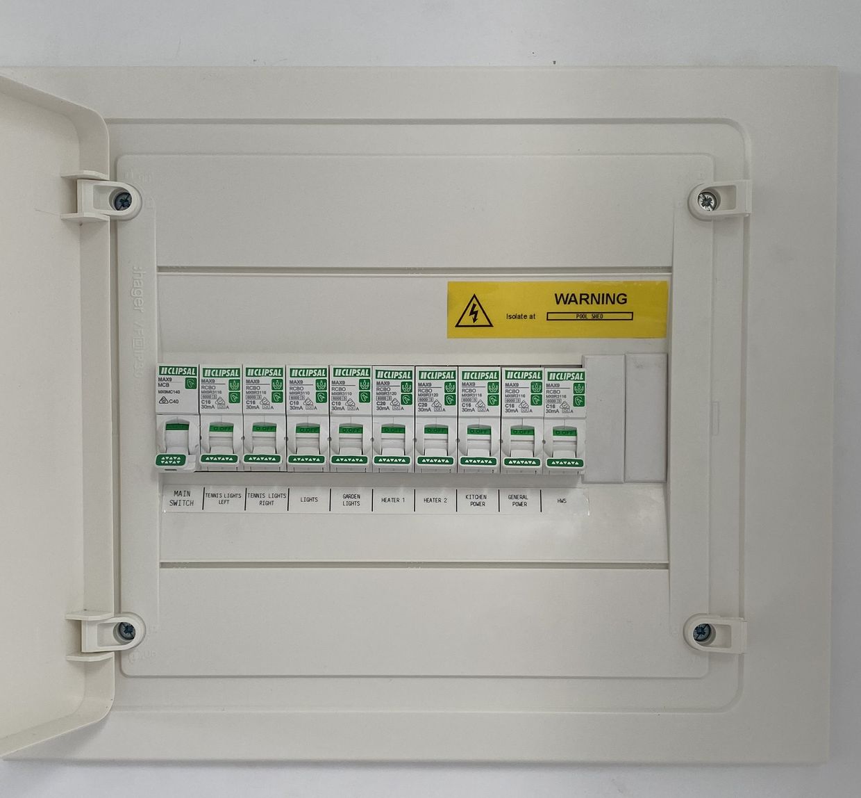 The same board finished, cover fitted and door open, every circuit labelled in plain words underneath its device: main switch, tennis lights left, tennis lights right, lights, garden lights, heater 1, heater 2, kitchen power, general power and hot water